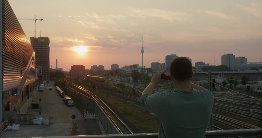 A man stands on an overpass over train tracks and takes a photo of the city with his smart phone
