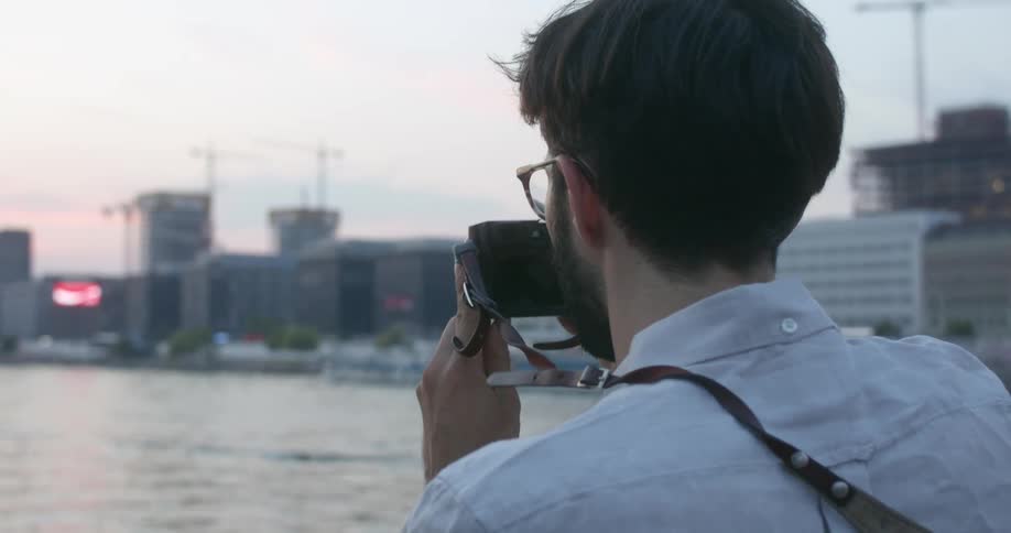 Young bearded man taking pictures of the sunset over the river in the city