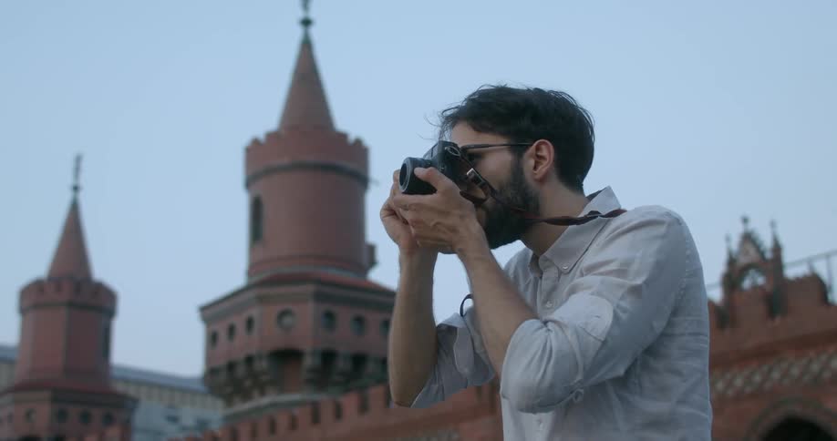 A man with glasses and a camera taking pictures with a building the sky and clouds in the background