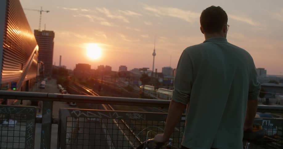 Man standing on a pedestrian overpass looks at the trains coming out of a station in the city at sunset