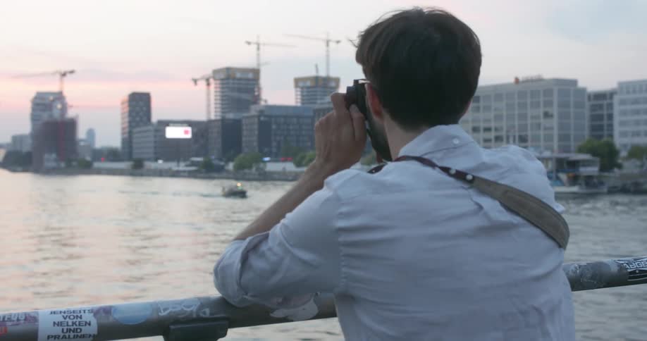 A man with a beard and glasses stands at a railing overlooking a body of water and a city while taking a picture with his camera