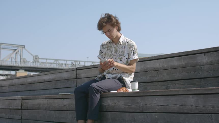 Young man sitting on a wooden bench outdoors picks up a phone