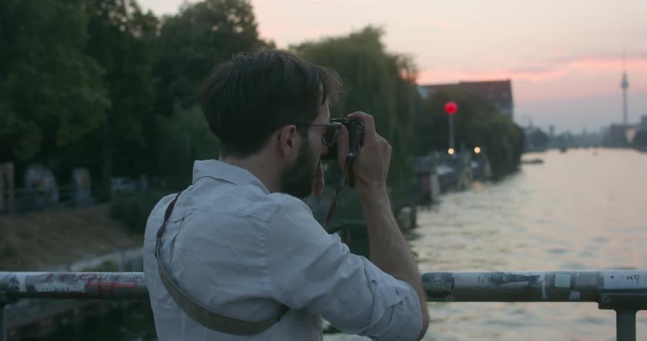 A man with a camera standing on a bridge over a river taking pictures with buildings the sky and clouds in the background