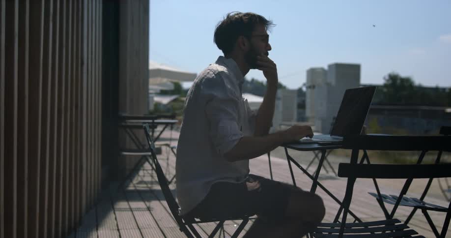 A man sits lost in thought in front of a laptop at a small table outdoors before checking his phone