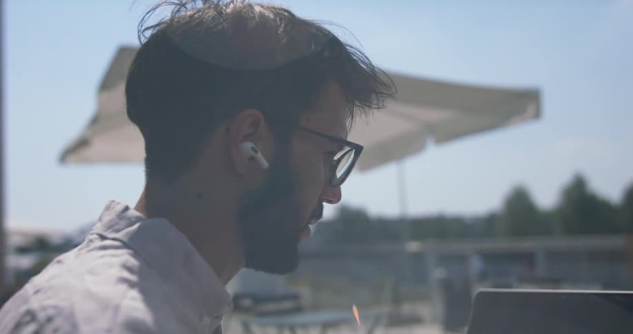 A man sitting at a small outdoor day with a laptop and smartphone retrieves a pair of ear buds from their holding case and places them in his ears