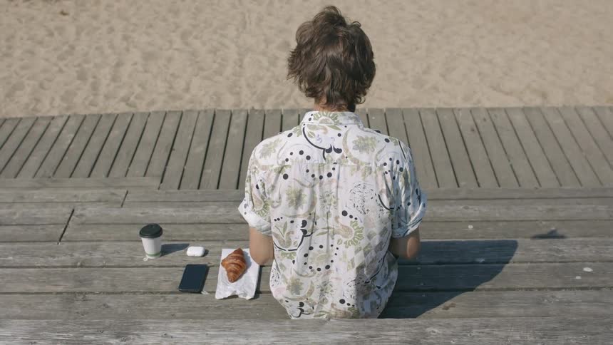 A teenage boy sits outside with a cup of coffee a croissant and his cell phone next to him