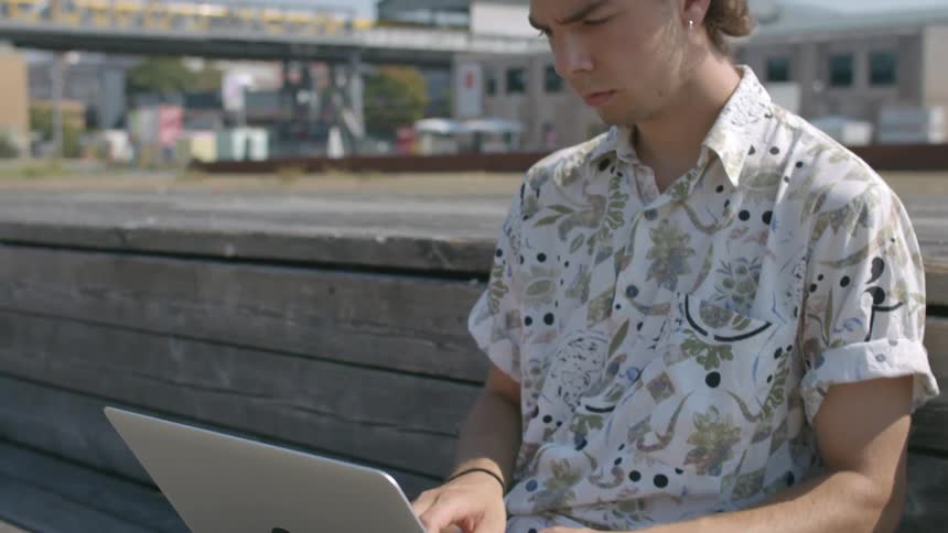A teenage boy types on his laptop outside as a train drives by in the distance