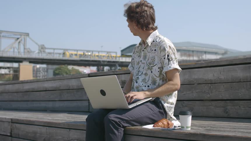 A young man sits outdoors on a wooden bench with his laptop computer