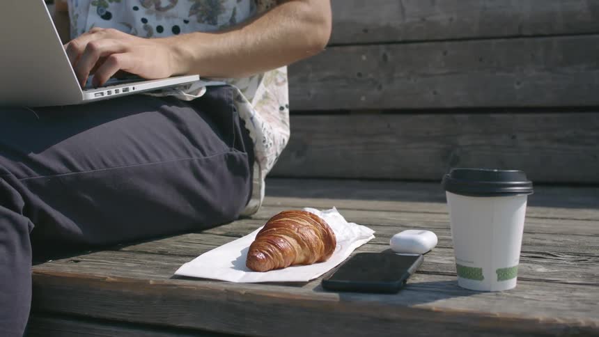 A man sits outside and types on his laptop with a croissant and cup of coffee and his cell phone and ear buds next to him