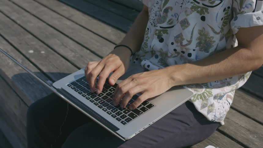 Man sits on bench typing on a laptop with coffee a croissant and a phone beside him