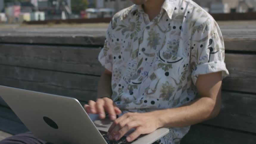 A man leans against a wall and checks his phone before continuing to work on his laptop