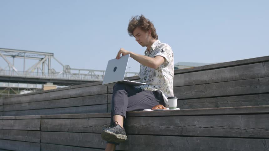 A man sitting outside on a bench opens a laptop and has a coffee and baked good next to him