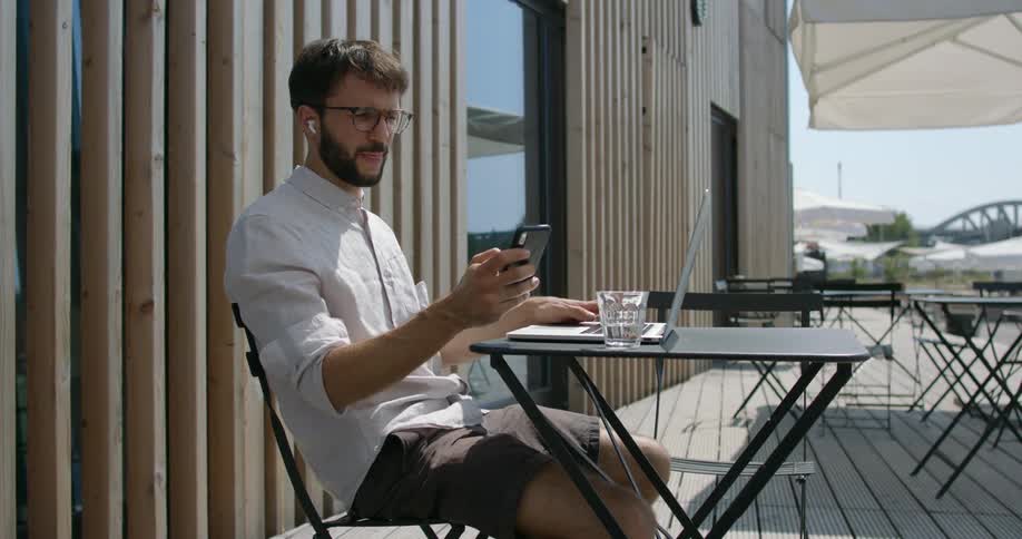 Man working on a laptop in an outdoors cafe checks his smartphone