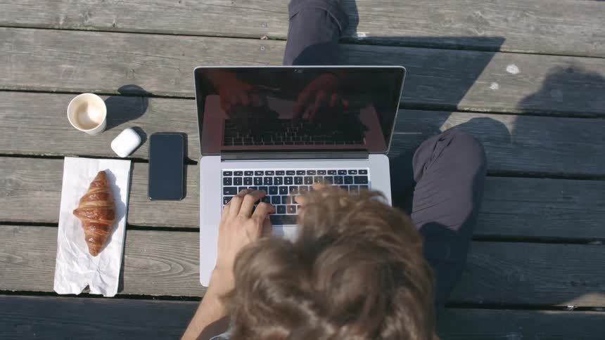 A man sits outdoors in the sun typing on his laptop with a croissant and cell phone besides him
