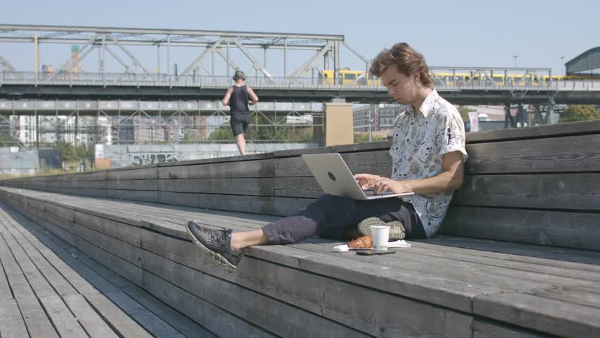 A man is sitting outdoors while typing on a laptop