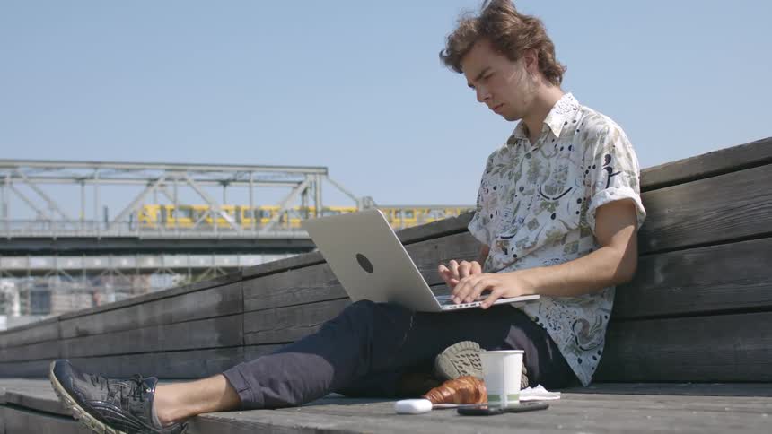 A teenage boy sits outside and uses his laptop as a train passes by behind him