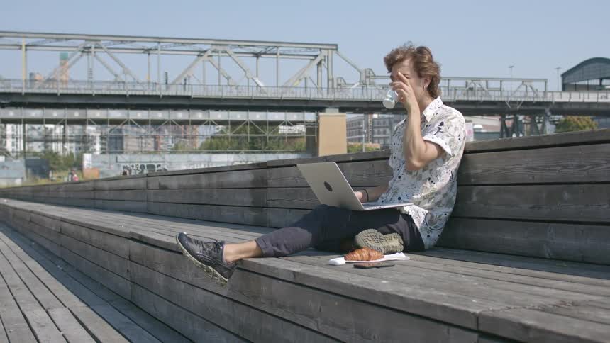 Man sits outside working on laptop while taking a drink as train passes over nearby bridge and pedestrians walk along