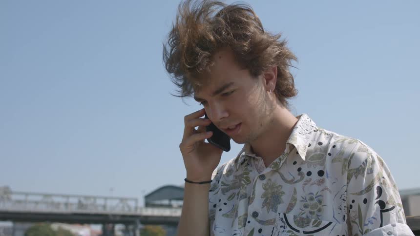 A male is sitting outdoors and talking on a cell phone while using a laptop