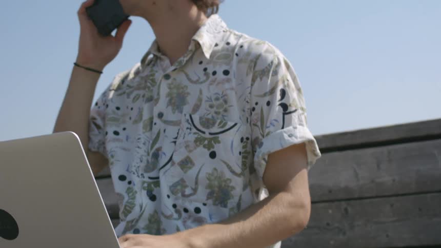 A young man sits on an outdoor bench with his cell phone and laptop