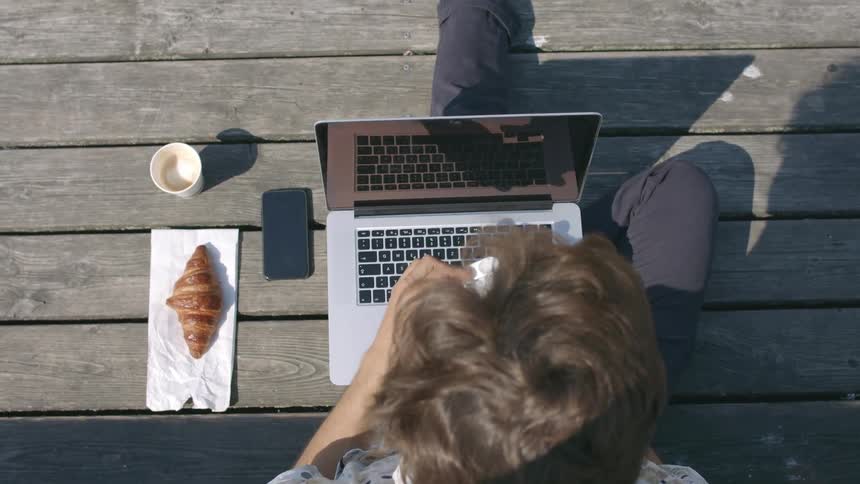 A man sitting on wood planks with a laptop cell phone croissant and coffee cup place ear buds in his ears