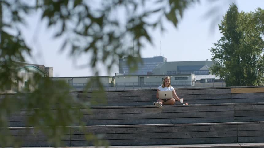 A woman sitting on bleachers typing on a laptop with buildings the sky and clouds in the background