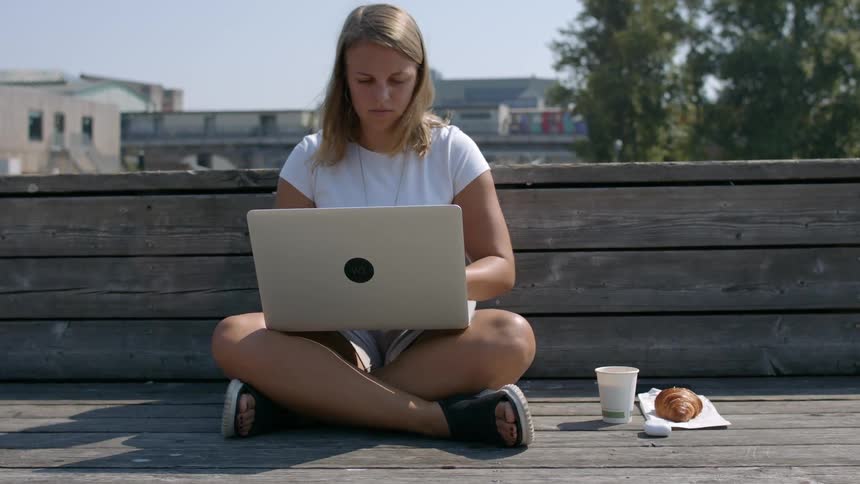 A white female sits on an outdoor bench while working on a laptop