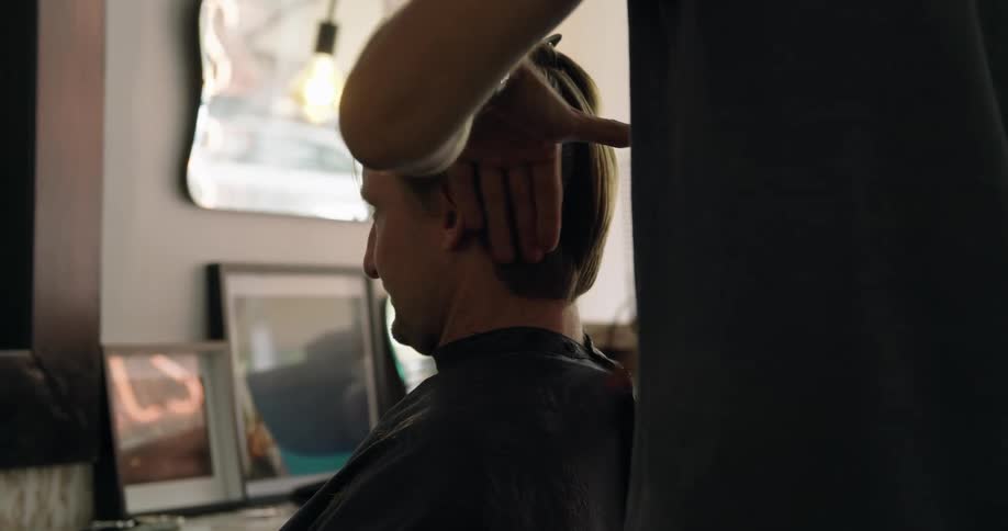 A man wearing a barber apron sittings in a chair in a barber shop as a stylist combs his hair
