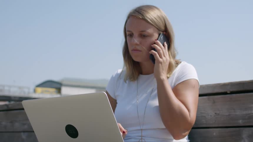 Young woman sitting outdoors working on a laptop picks up a phone to answer a call