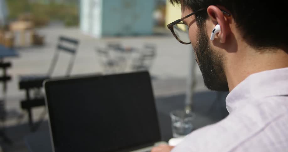 A bearded man with glasses and ear buds sits outdoors at a table typing on a laptop during a sunny day