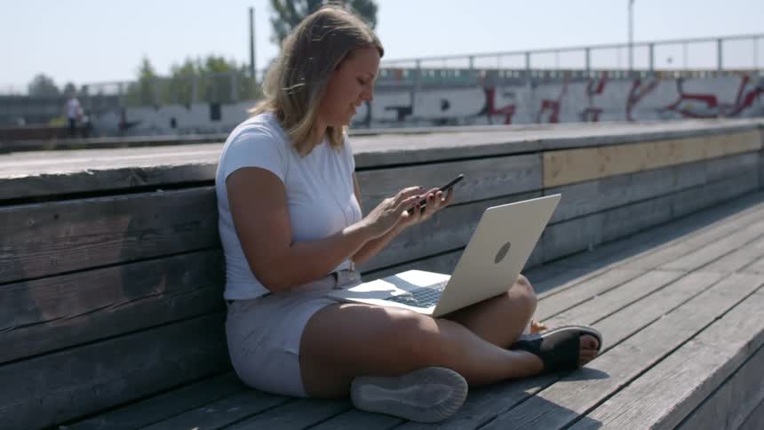 Woman sitting on a bench in a park talking on her phone while on her laptop