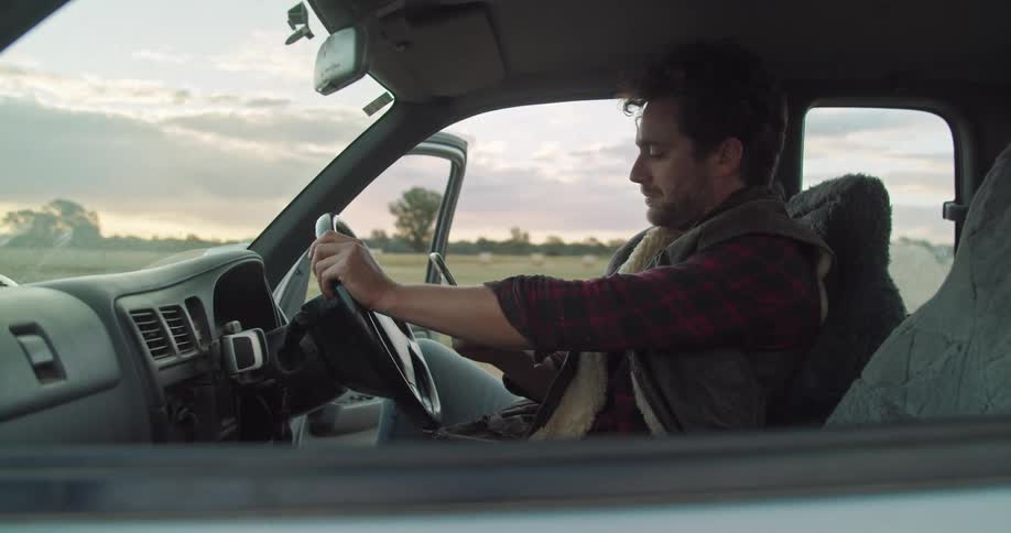Man sitting in a car texting on a smartphone in a rural field at sunset