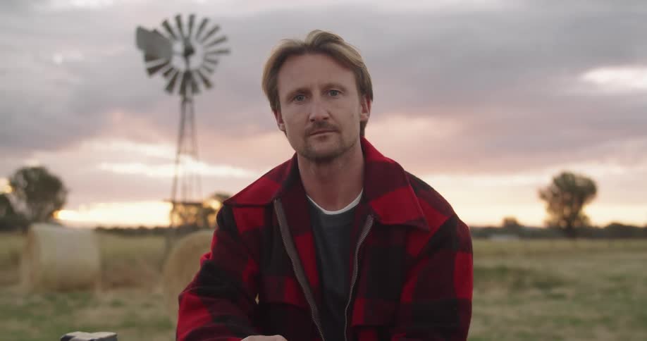Farmer sitting in front of a wind mill in a farm at sunset looking at the camera