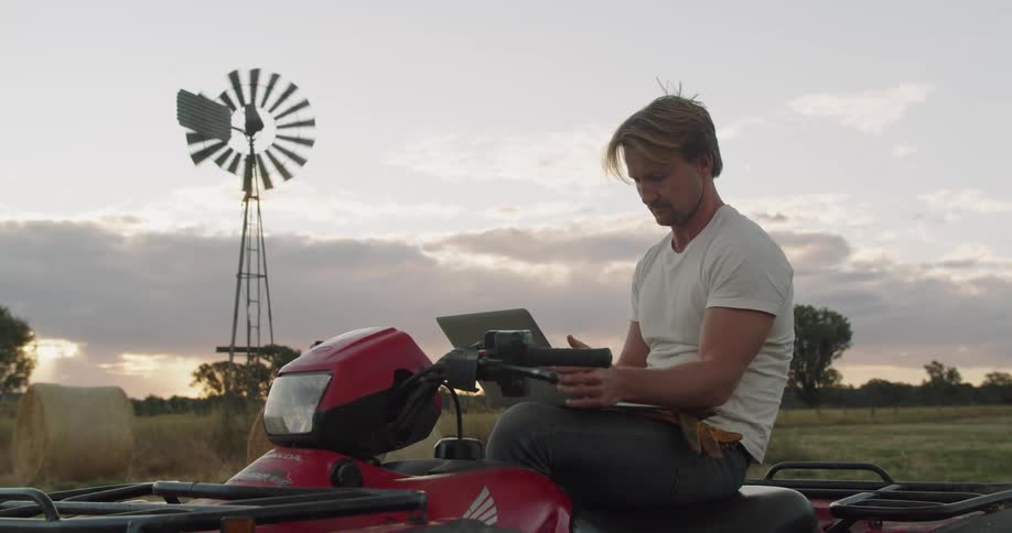 A man sits on a four wheeler on a farm holding a laptop