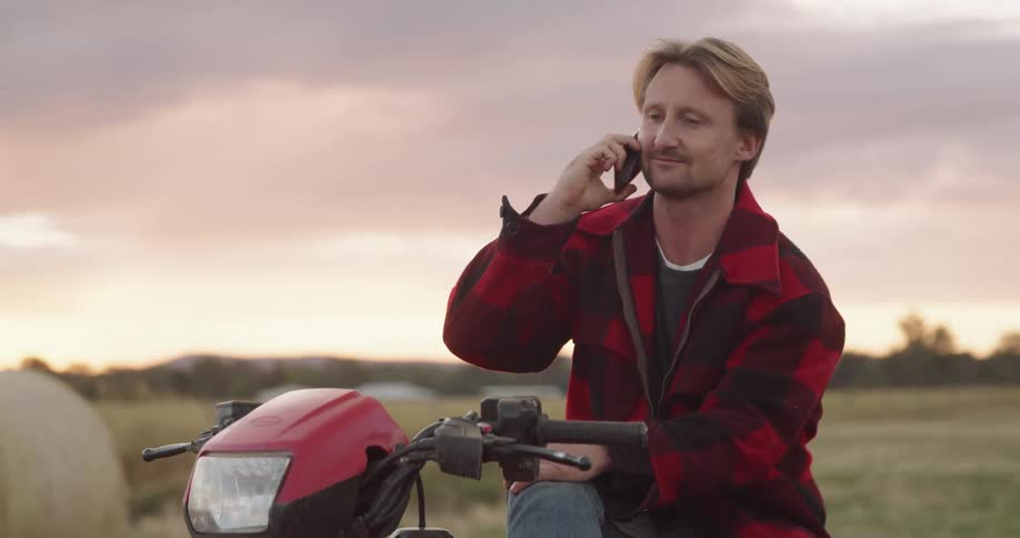 Man sits on vehicle in field near hale bays talking on mobile phone under a red sky
