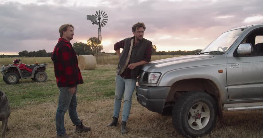 One man leaning against a silver car talking to another man outside next to a farm field at sunset