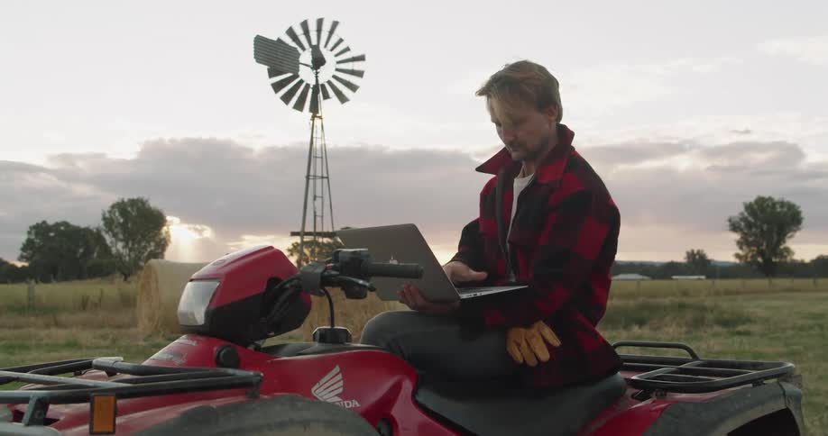A man sits on a four wheeler in a farm typing on a laptop