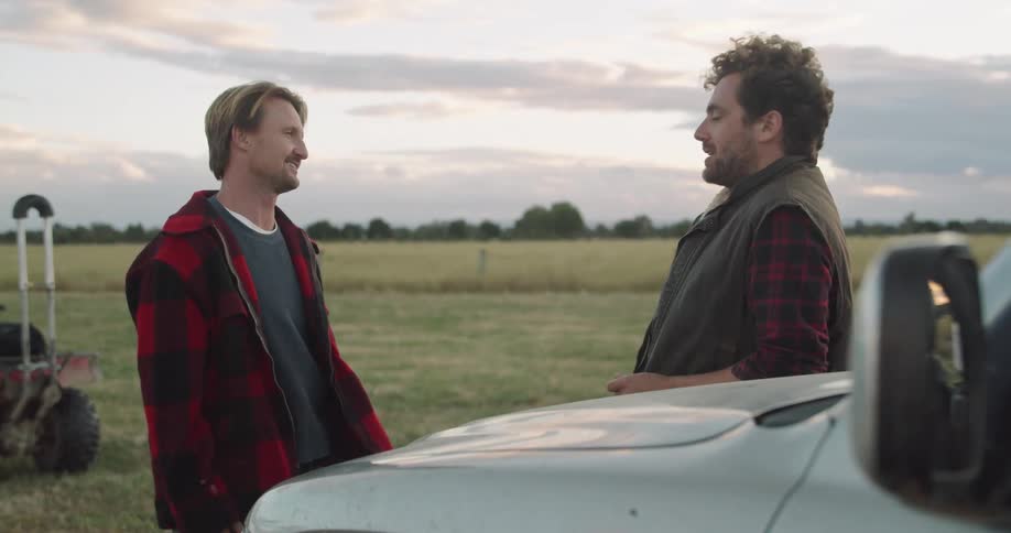A pair of men speak with one another while standing near a parked vehicle in a field in the evening