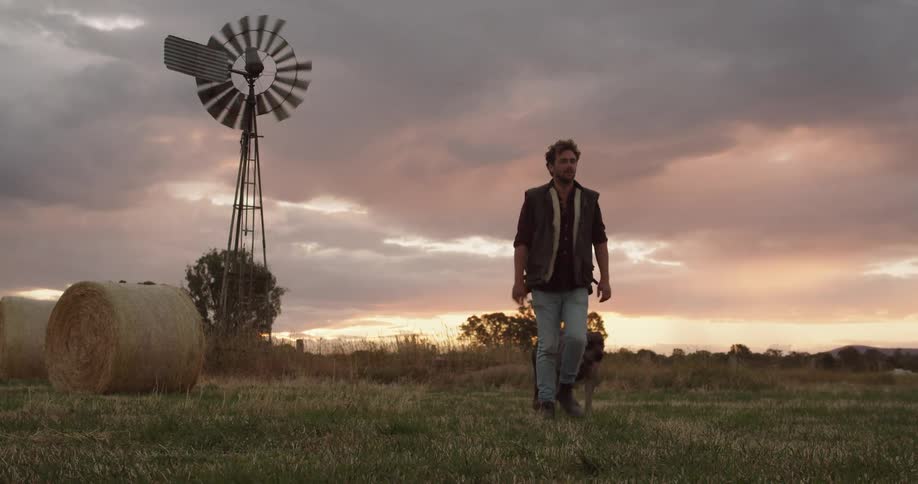 Man walking out in a field next to a windmill and a haystack with his pet dog at sunset