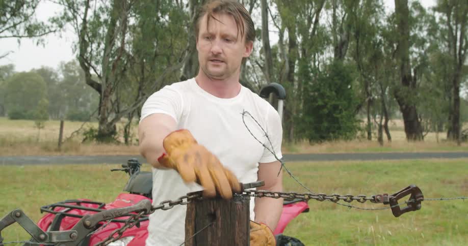 A man steps off of his vehicle a walks over to a fence post too pick up a tool atop it