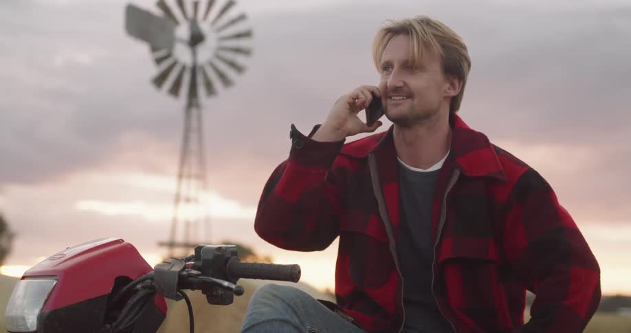 A man wearing a red plaid jacket sits on a motorcycle talking on a cell phone in a rural area