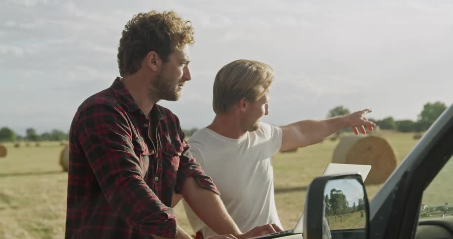 Two men stand outside a truck in a rural area looking at a laptop of the hood of the truck and talking