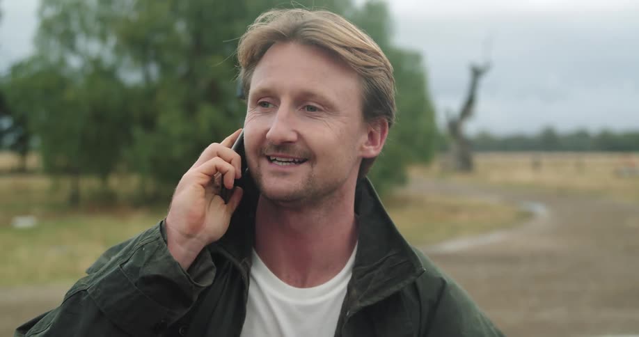 A man standing and talking on a phone with trees the sky and clouds in the background