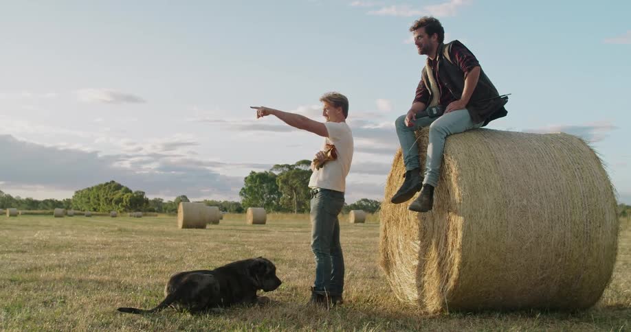 A dog lies at a mans feet as he points across a farm field and talks with a man sitting on a hay bale