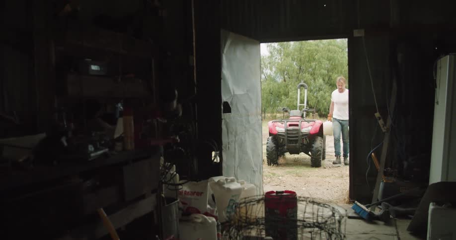 A man gets off of a four wheeler and carries two gas jugs into a barn