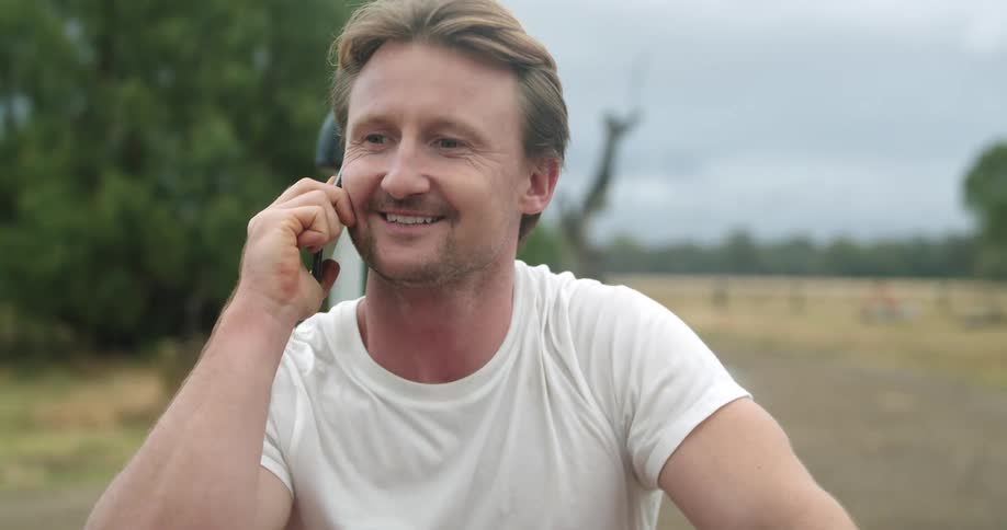 Man riding a motorcycle parked on a field outdoors while talking on a smartphone