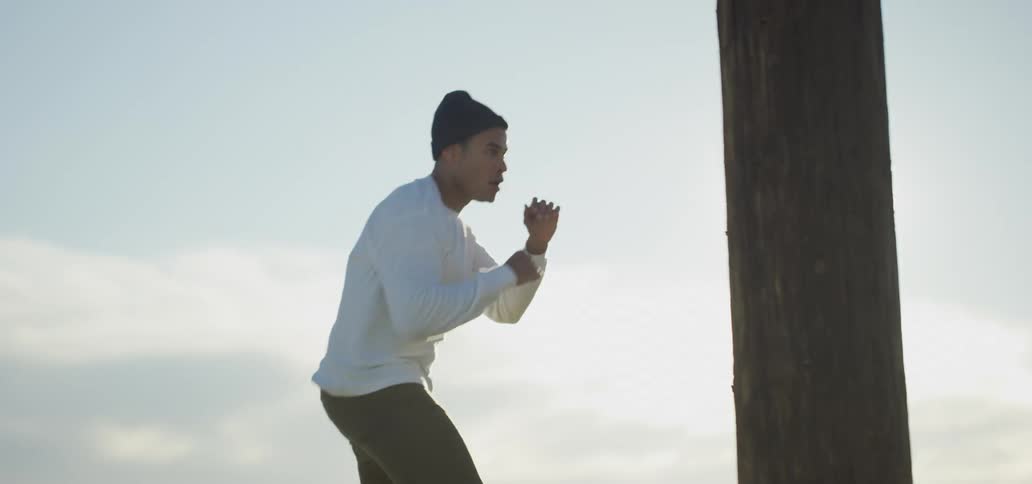 A young man shadow boxes while facing a utility pole outdoors on a sunny day
