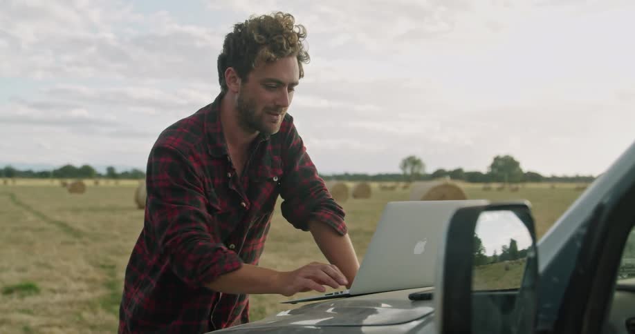 A man typing on a laptop on the hood of a vehicle with a grassy field the sky and clouds in the background
