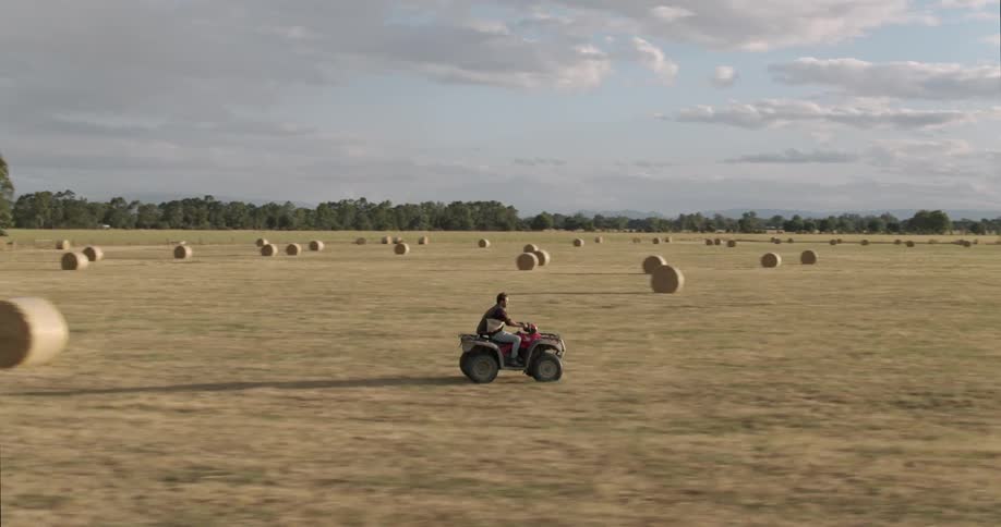 Man drivng an all terrain vehicle on a field filled with hay bales at sunset