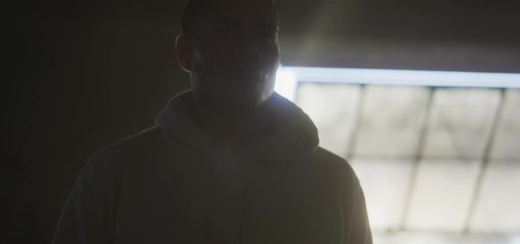 A man wearing a hoodie sweatshirt walks in a building with windows in shadows with a brief beam of sunlight