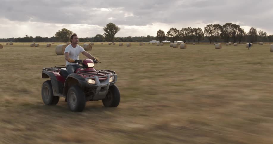 A man rides an all terrain vehicle through a grassland filled with hay bales on a cloudy evening